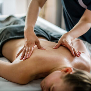 Eine Person liegt entspannt auf einer Massageliege, während eine andere Person ihr mit beiden Händen den Rücken massiert. Die liegende Person ist mit einem dunklen Tuch bedeckt und die Szene wirkt ruhig und wohltuend.