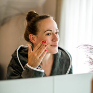 Eine Frau im Bademantel steht vor einem Spiegel und trägt mit einem Lächeln Gesichtscreme auf ihre Wange auf. Sie hat ihre Haare zu einem Dutt gebunden und ihre Fingernägel sind rot lackiert. Im Hintergrund fällt weiches Tageslicht durch ein Fenster – die Szene vermittelt Ruhe und Selbstfürsorge.