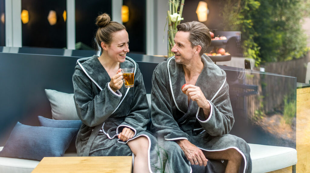 Frau und Mann im Bademantel schauen sich draußen auf einer Liege an und lächeln