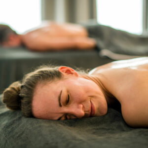 Eine Frau liegt entspannt mit geschlossenem Auge und einem zufriedenen Lächeln auf einer Massageliege, zugedeckt mit einem dunklen Handtuch. Im Hintergrund ist eine weitere Person unscharf zu erkennen, ebenfalls liegend. Die Atmosphäre wirkt ruhig und wohltuend – typisch für eine Wellness- oder Spa-Anwendung.