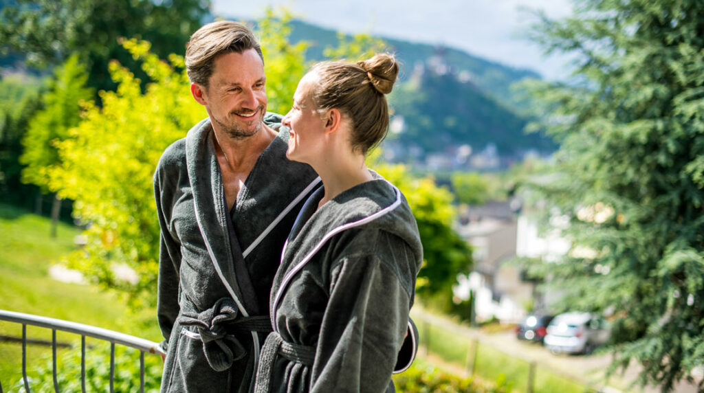 Paar im Bademantel steht vor Wiese und Bäumen und lacht zufrieden